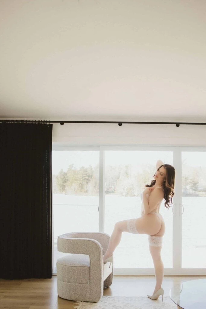 Ava Marie, arrayed in refined white lingerie and lofty heels, is thoughtfully setting a pose by a window that overlooks the outdoors. She is comfortably immersed in the soft, natural light streaming in. Ava Marie Halifax's Elite Independent Companion
